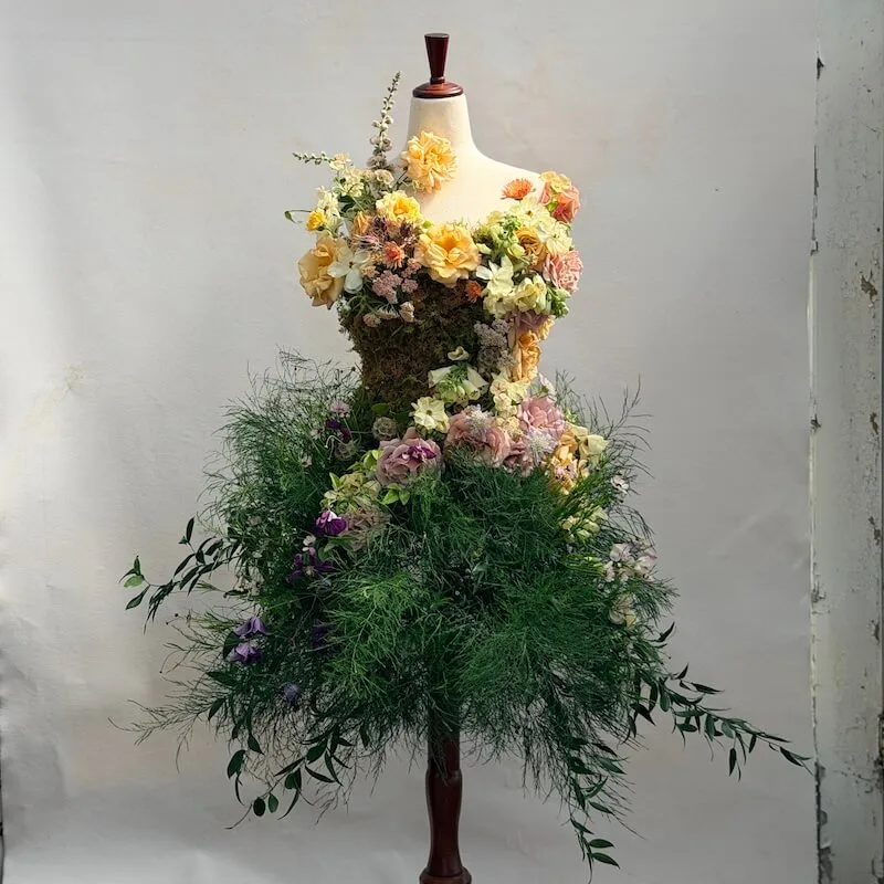 Floral dress art-piece — a dress form gown with peach, yellow, and blush roses on the bodice and a lush green fern skirt with hydrangea accents
