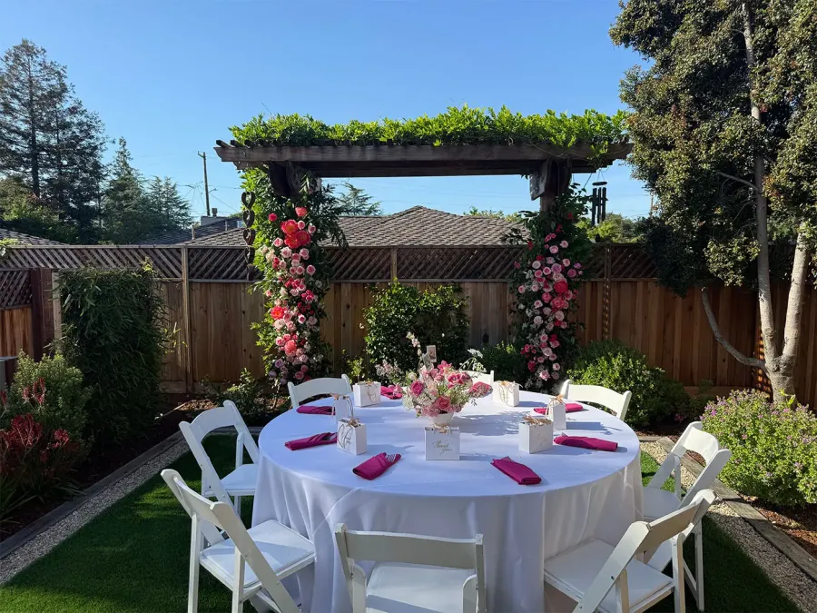 Pink and red rose floral arch over a round table with white linens and fuchsia napkins in a garden setting by Tracy Catering Company